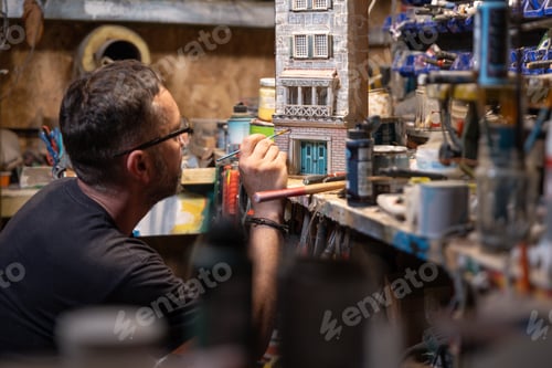 Preview: Modeller man working at scale model of miniature building in his workshop full of tools