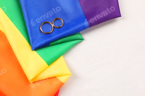 Preview: Rainbow LGBT flag and wedding rings on white background, top view. Space for text
