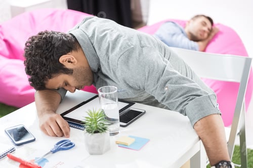 Preview: Young businessmen sleeping on the table and poufes at modern office, business teamwork