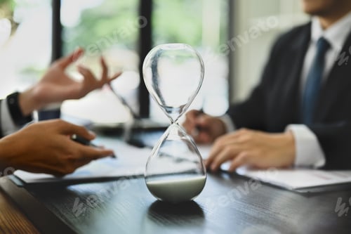 Preview: Closeup hourglass on meeting table with blurred two businesspeople working on background