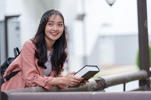 Preview: Happy young asian woman with backpack holding a book and smiling while leaning on a railing in a uni