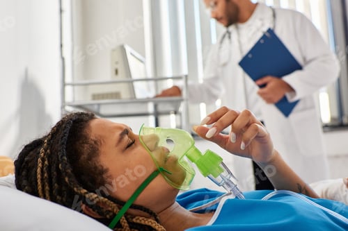 Preview: focus on african american woman lying in bed with oxygen mask with blurred indian doctor on backdrop