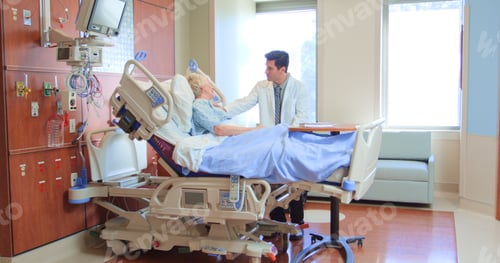 Preview: Male Doctor Wearing White Coat Visiting Senior Female Patient In Hospital Bed Giving Her Good News