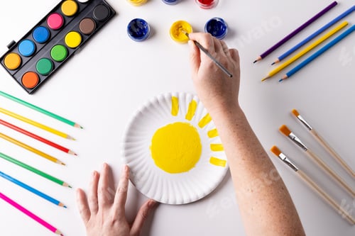 Preview: Overhead of hands painting with yellow paint on paper plate, with art materials on table top