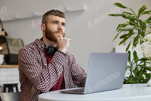 Preview: Happy young attractive ginger bearded man working at a laptop while sitting in a cafe