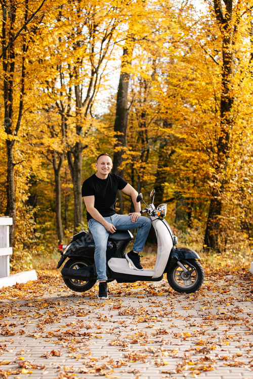 Preview: Smiling young man sitting on retro white scooter in autumn forest or park with trees with yellow