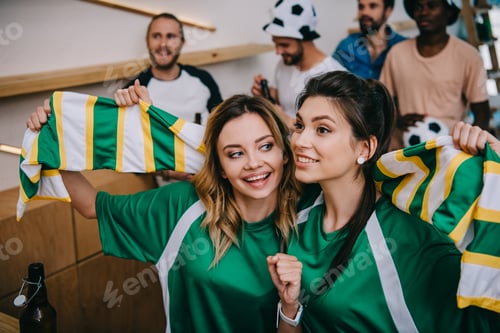 Preview: women in green fan t-shirts holding fan scarf and their male friends standing behind during watch of