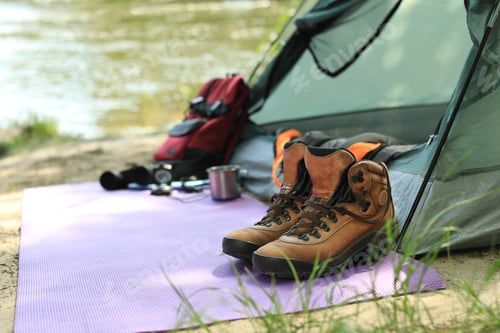 Preview: Boots and camping equipment near tent on riverbank. Space for text