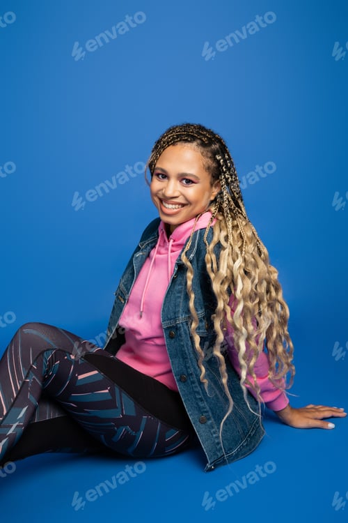 Preview: happy african american woman with long hair looking at camera, sitting on blue background, sporty