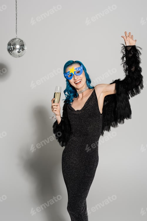 Preview: joyful woman in party mask and black faux fur jacket holding champagne and waving hand on grey