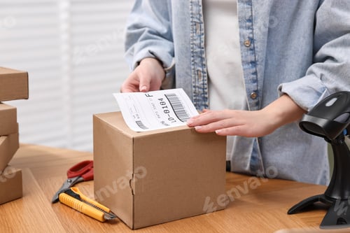 Preview: Parcel packing. Post office worker sticking barcode on box at wooden table indoors, closeup