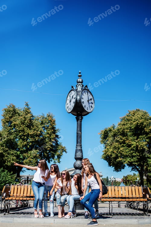 Preview: Six beautiful young girls sitting on a bench next to the old street clock in the park.