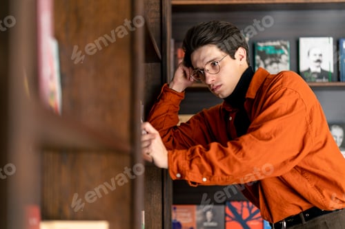 Preview: young guy in glasses stands near the bookshelves thoughtfully chooses a book in university library