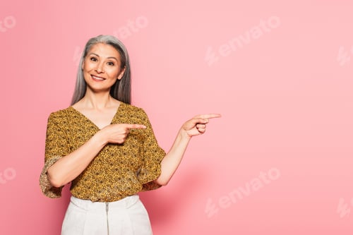 Preview: positive asian woman pointing with fingers while smiling at camera on pink background