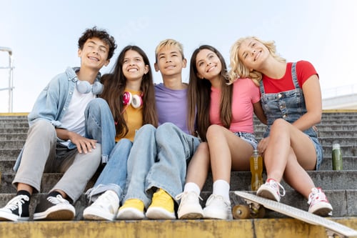 Preview: Group of smiling friends, multiracial teenagers wearing colorful t shirts hugging, sitting on stairs