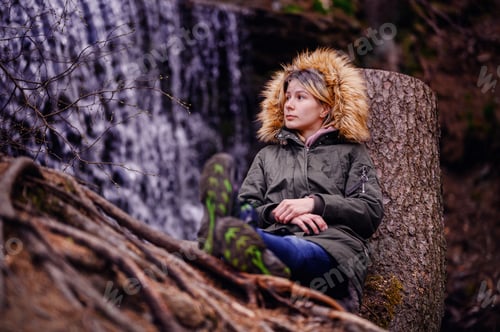 Preview: Young woman sits leaning on a stump in green parka with fur before waterfall alone in forest