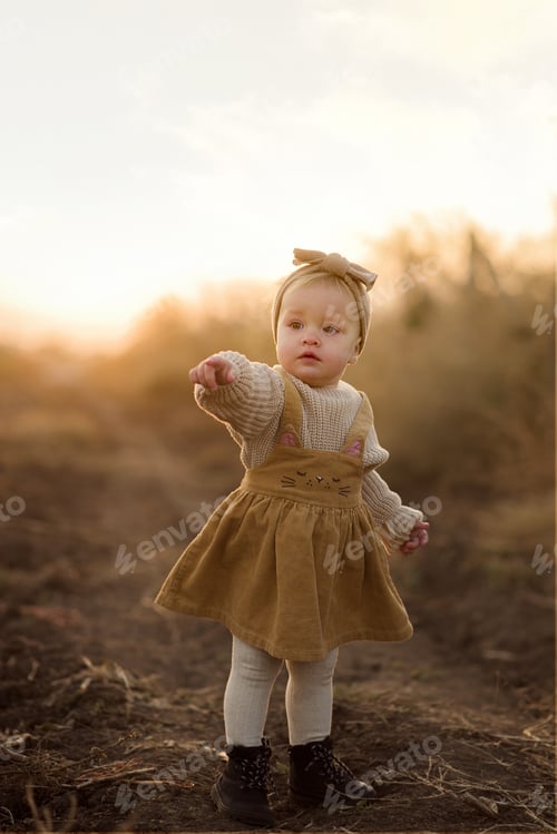 Preview: Little pretty girl is standing and showing something with her finger in autumn field at sunset