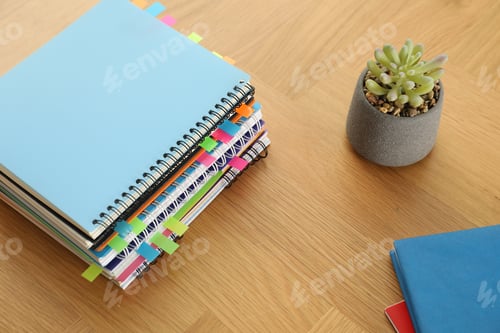 Preview: Stack of notebooks with colorful bookmarks and houseplant on wooden table, above view