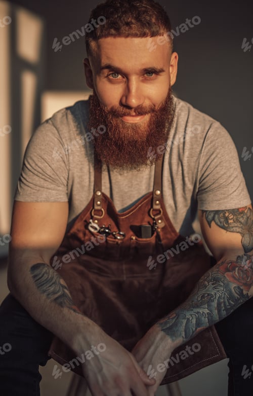 Preview: Confident bearded hairdresser in working apron sitting leaning on knees