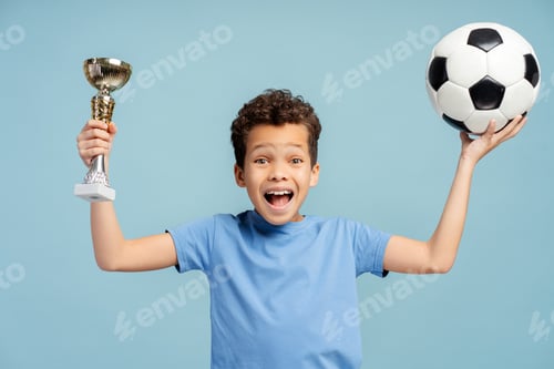 Preview: Little footballer holding a golden trophy, isolated on blue background. Playing football concept