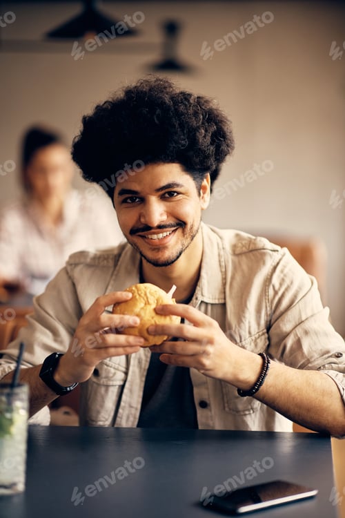 Preview: Young happy Muslim man eating burger in cafeteria and looking at camera.