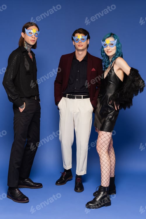 Preview: full length of stylish queer friends in party masks looking at camera while standing on blue