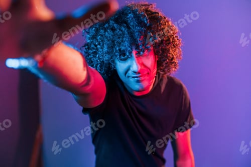 Preview: Takes selfie. Young beautiful man with curly hair is indoors in the studio with neon lighting