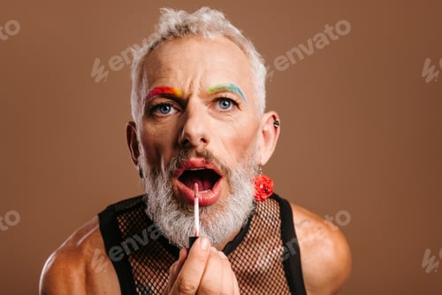 Preview: Bearded mature gay man with rainbow colored eyebrows applying lipstick against brown background