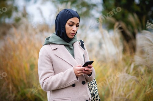 Preview: Portrait of young muslim woman in coat, standing outdoor in city park, scrolling smartphone.