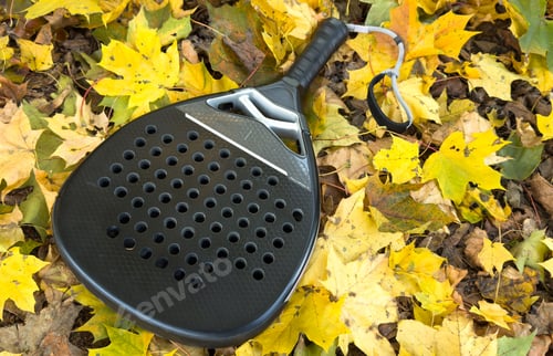 Preview: Top view of a black padel racket on a green court grass turf behind the net outdoors. Paddle is a
