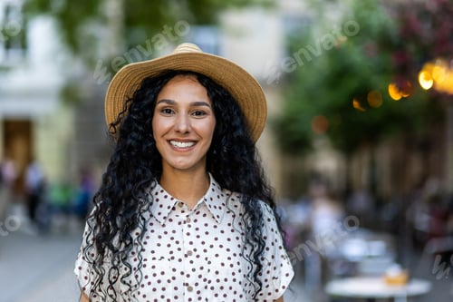Preview: Beautiful young Latin American woman portrait, woman walking in evening city in hat with curly hair