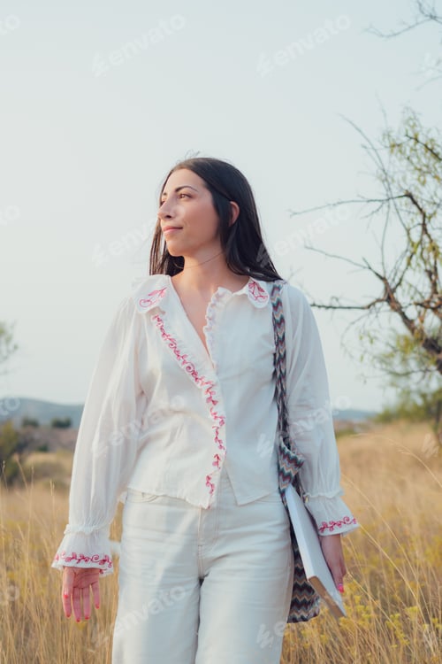 Preview: Young woman dressed in white walks through a meadow at sunset