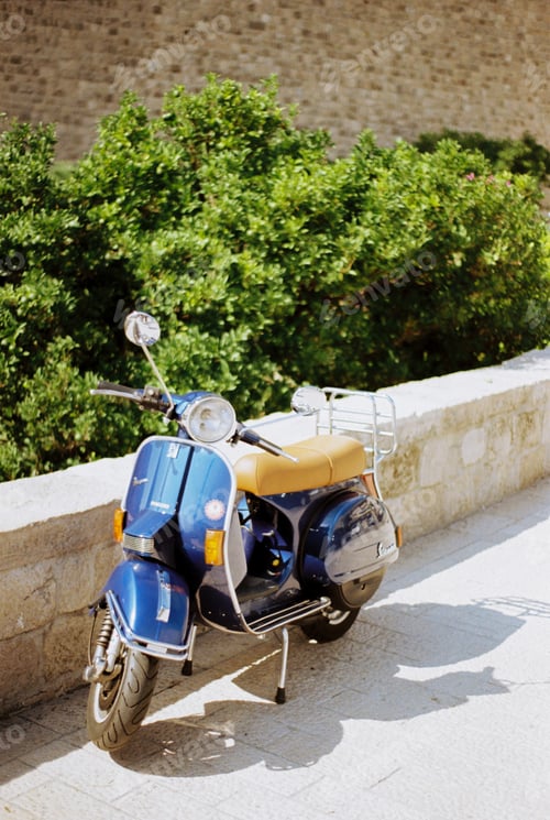 Preview: Blue motor scooter stands on paving stones near a stone fence