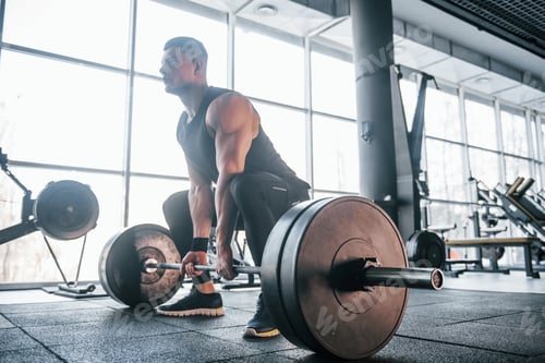 Preview: Strong young man in sportive clothes doing exercises with heavy weights in the gym