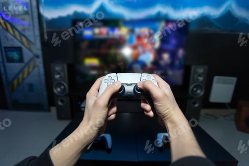 Preview: Close up of man hands holding wireless joysticks on foreground. Modern TV set with console
