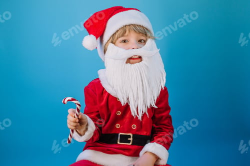 Preview: Funny boy in Santa Claus hat and sweet caramel cane on blue studio background