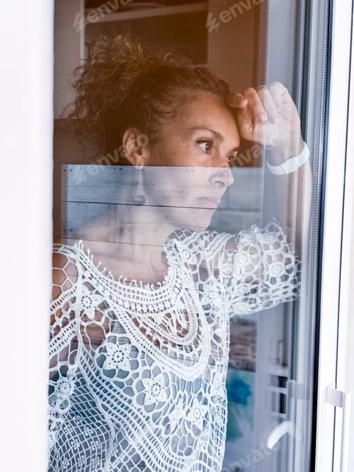 Preview: Sad adult woman inside home looking outside through the door window glass.