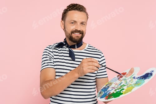 Preview: positive french artist with neck scarf holding palette with colorful paints and paintbrush isolated