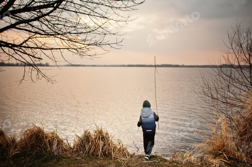 Preview: Boy in hoodie by the lake with a reed in his hands.