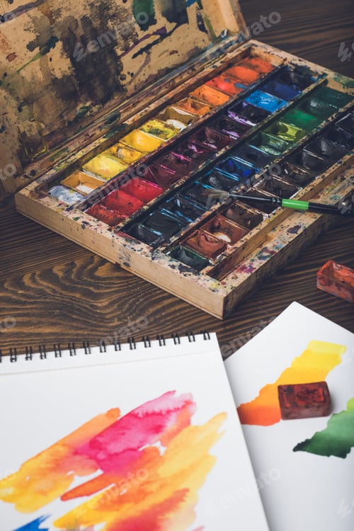 Preview: close-up view of watercolor paints, paintbrush and abstract sketches at designer workplace