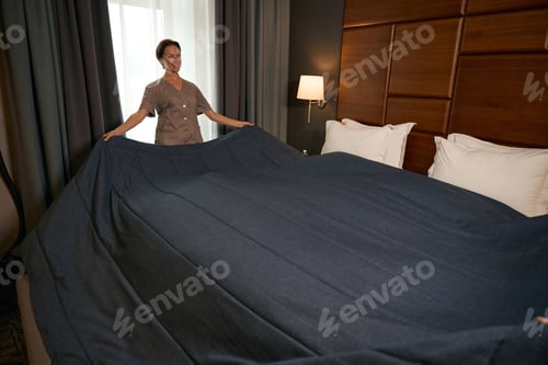 Preview: Joyful young chambermaid busy with bed-making procedure in guest suite