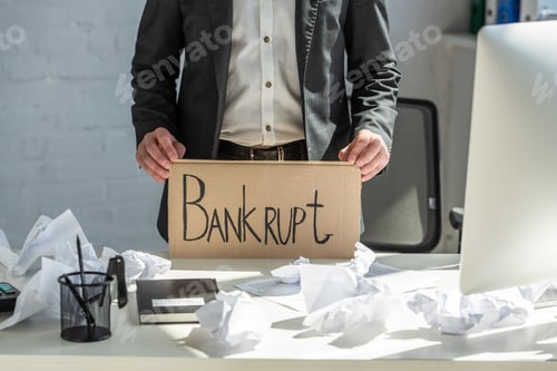 Preview: Cropped view of businessman holding cardboard with bankrupt lettering, while standing near table on