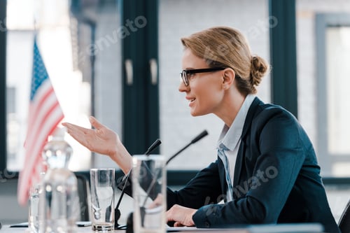 Preview: side view of beautiful diplomat in eyeglasses gesturing while talking near microphone and american