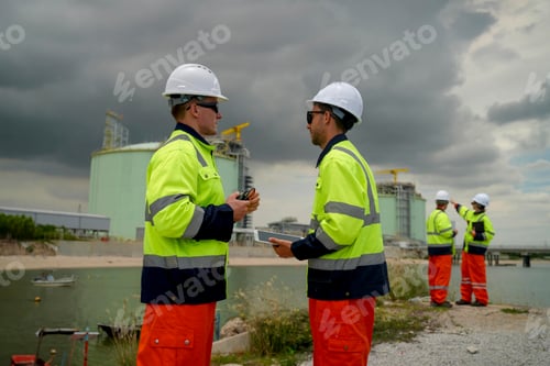 Preview: Workers in safety gear collaborate by the water near an industrial facility on a cloudy day