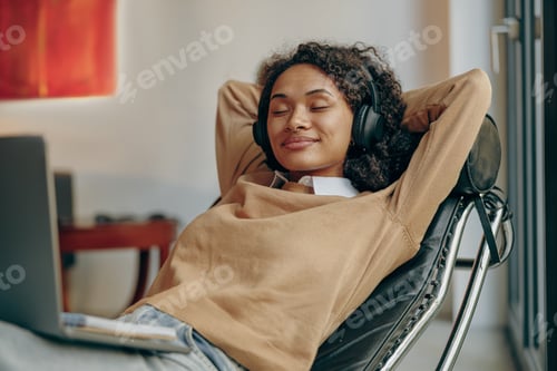 Preview: Smiling female freelancer in headphones is resting while lying on chair during recess at home