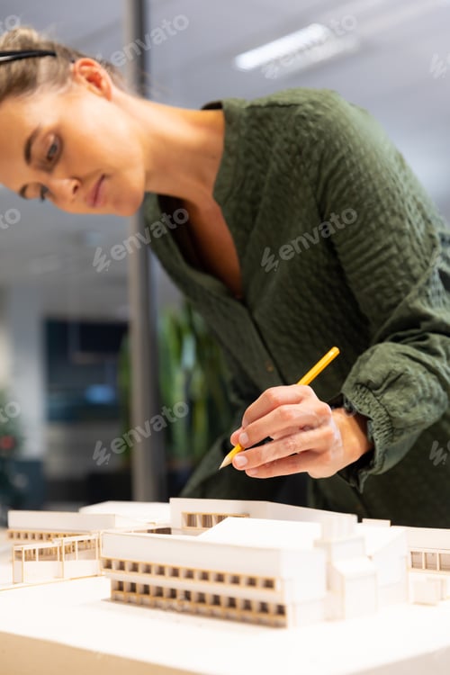 Preview: Caucasian female architect analyzing architectural model on desk with pencil at modern workplace