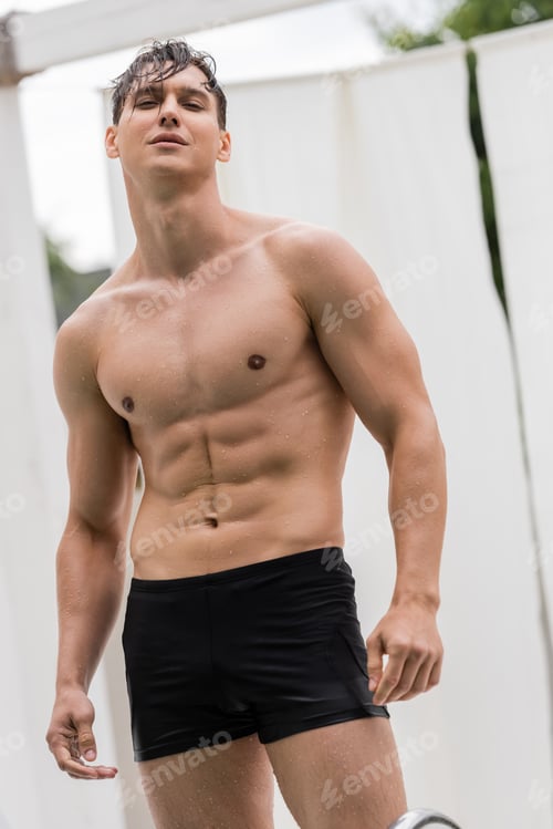 Preview: wet and muscular man in swimming trunks looking at camera outdoors
