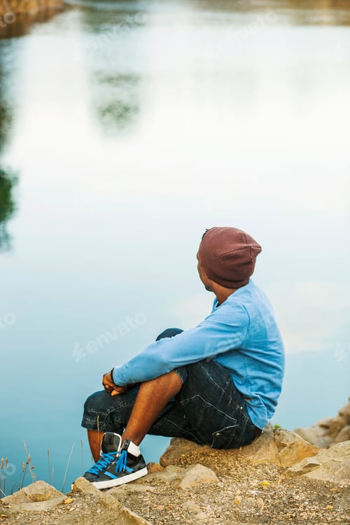 Preview: Side view of man sitting on rock by lake