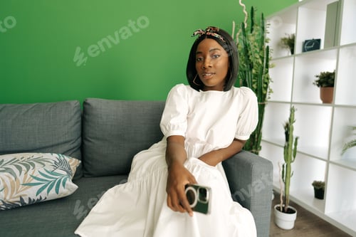 Preview: Young african woman sitting on couch at home and using smartphone