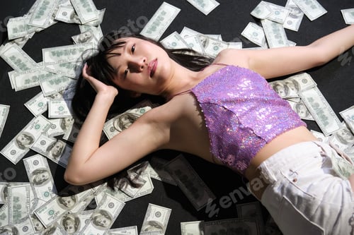 Preview: Young asian woman surrounded by dollar bills laying on the floor.